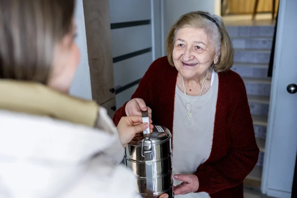 A GA Foods driver hands a stainless steel meal container to a smiling senior woman at her front door — meal delivery route optimization in action